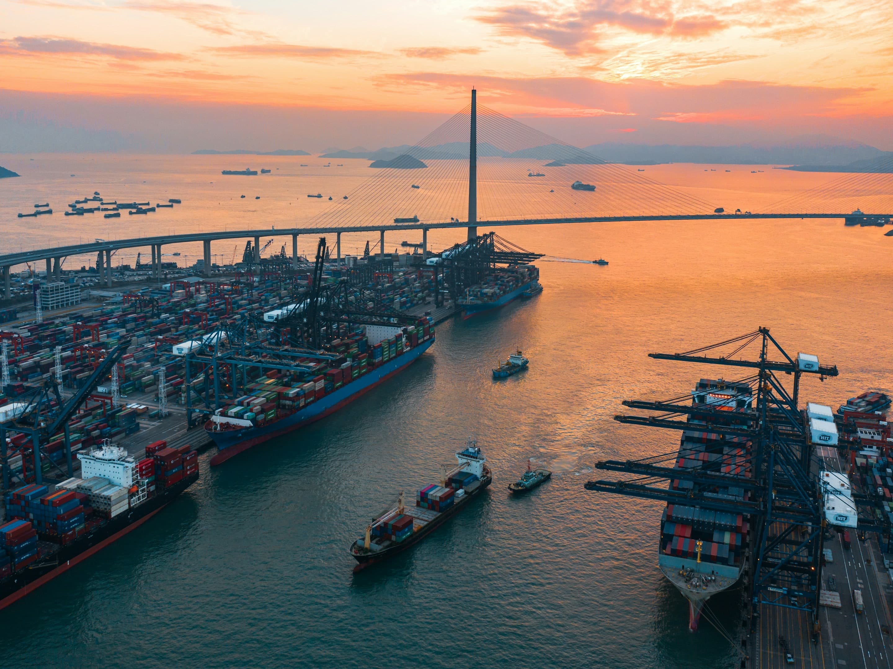 Terminal de contêineres de Hong Kong ao entardecer, com navios atracados e a ponte ao fundo.